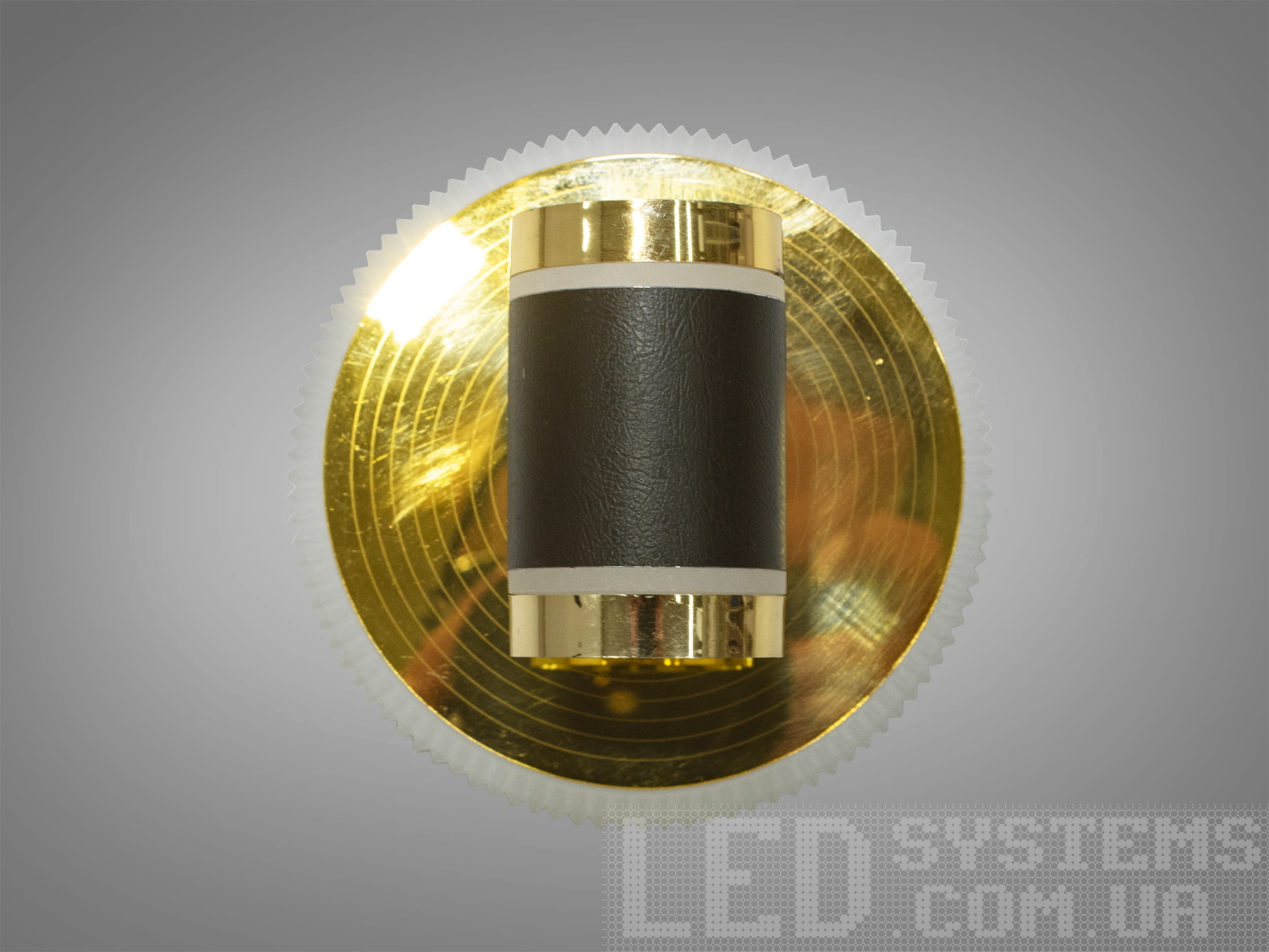 Компактний настінний світильник з золотистою круглою основою та циліндричним абажуром з чорної шкіри, ідеальний для локального освітлення.