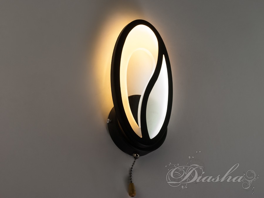 Настінний світильник овальної форми з чорним обрамленням, що створює теплу атмосферу, має вимикач на ланцюжку, підходить для спальні.