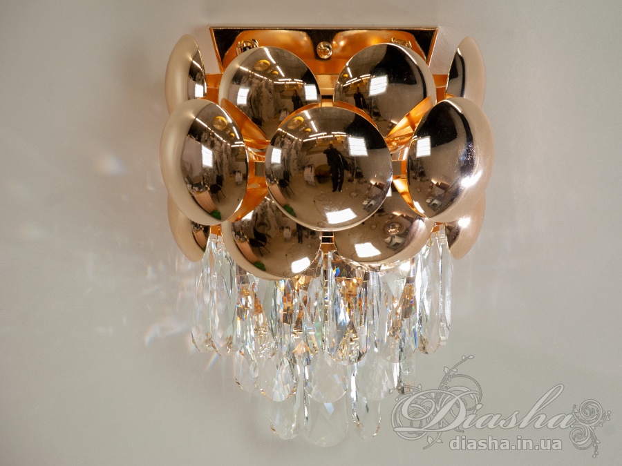 Золота металева основа світильника надає йому чарівності та тепла, що ідеально поєднується з прозорістю та блиском кришталів, які підвішені у каскаді під круглими випуклими золотистими елементами.
